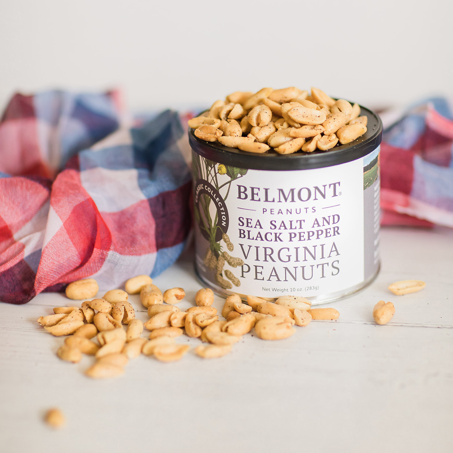 An open can of Sea Salt & Black Pepper gourmet peanuts spills onto a white surface beside a red, white, and blue plaid cloth.