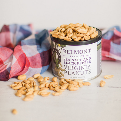 An open can of Sea Salt & Black Pepper gourmet peanuts spills onto a white surface beside a red, white, and blue plaid cloth.