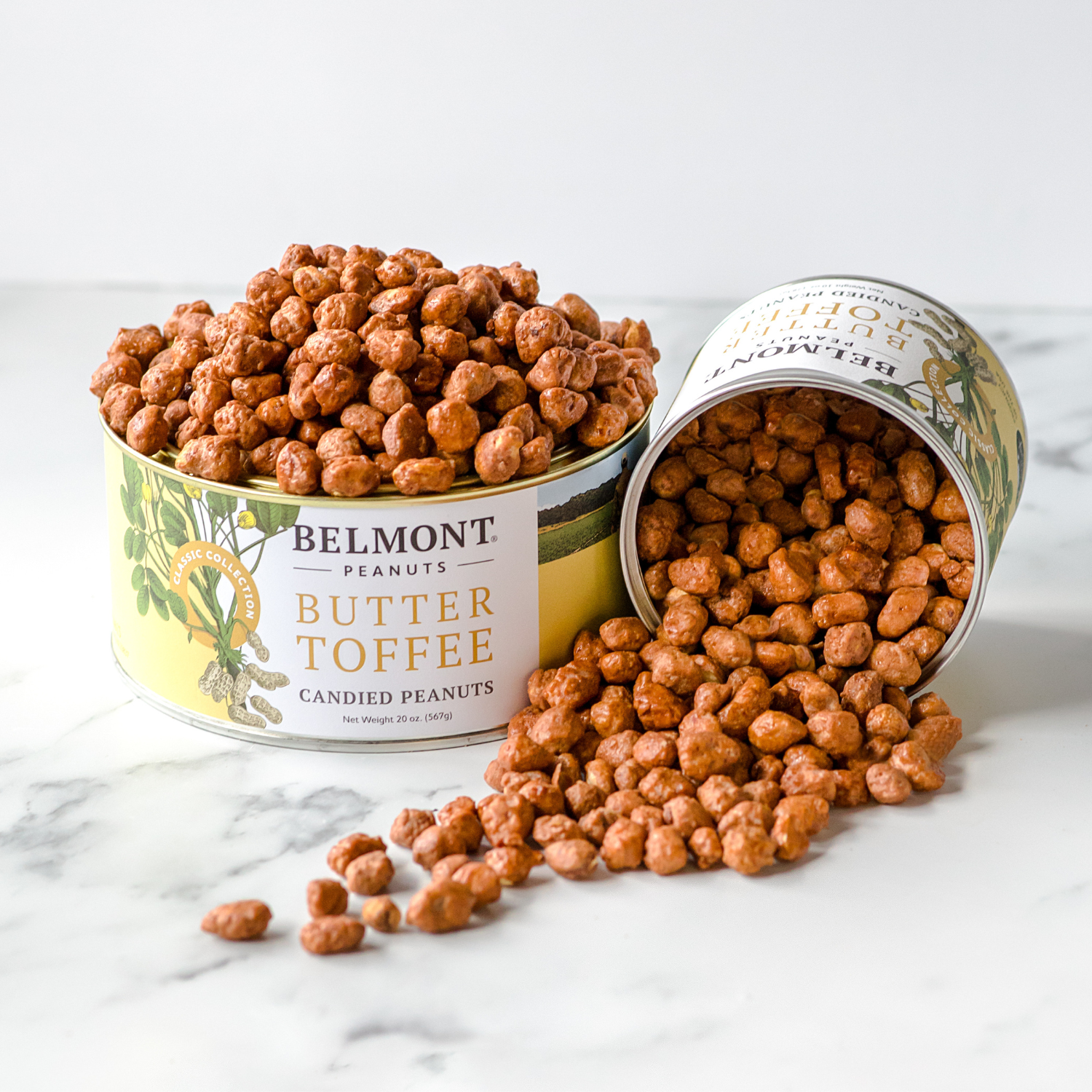 Two cans of Butter Toffee are displayed; one stands upright and full, while the other is tipped over with sweet candy-coated peanuts spilling out onto a white marble surface.