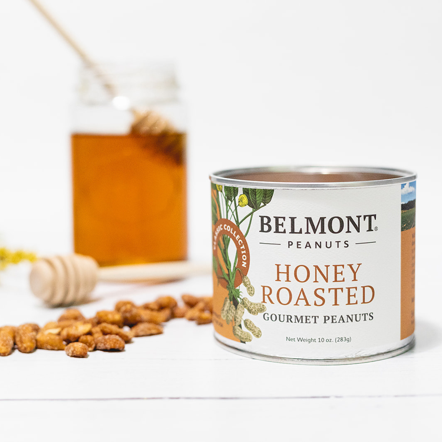 A can of Honey Roasted peanuts sits on a white surface with jumbo peanuts scattered nearby, a honey dipper, and a jar of honey in the background.