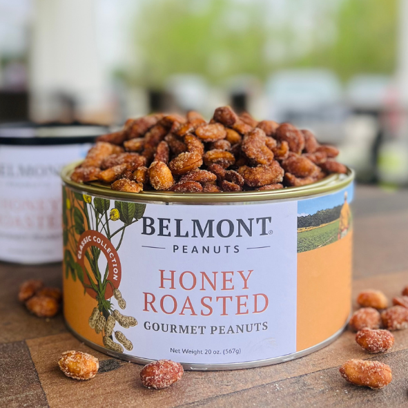 An open can of Honey Roasted peanuts displays sugared jumbo peanuts, some scattered on a wooden surface, with the vibrant label contrasting against a blurred outdoor background.