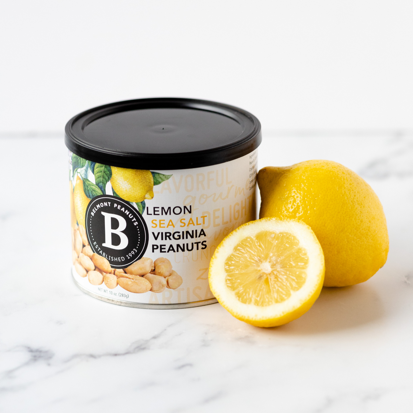 A can of Lemon Sea Salt peanuts rests on a white surface beside a whole lemon and a lemon half, with a label displaying peanut and lemon designs.