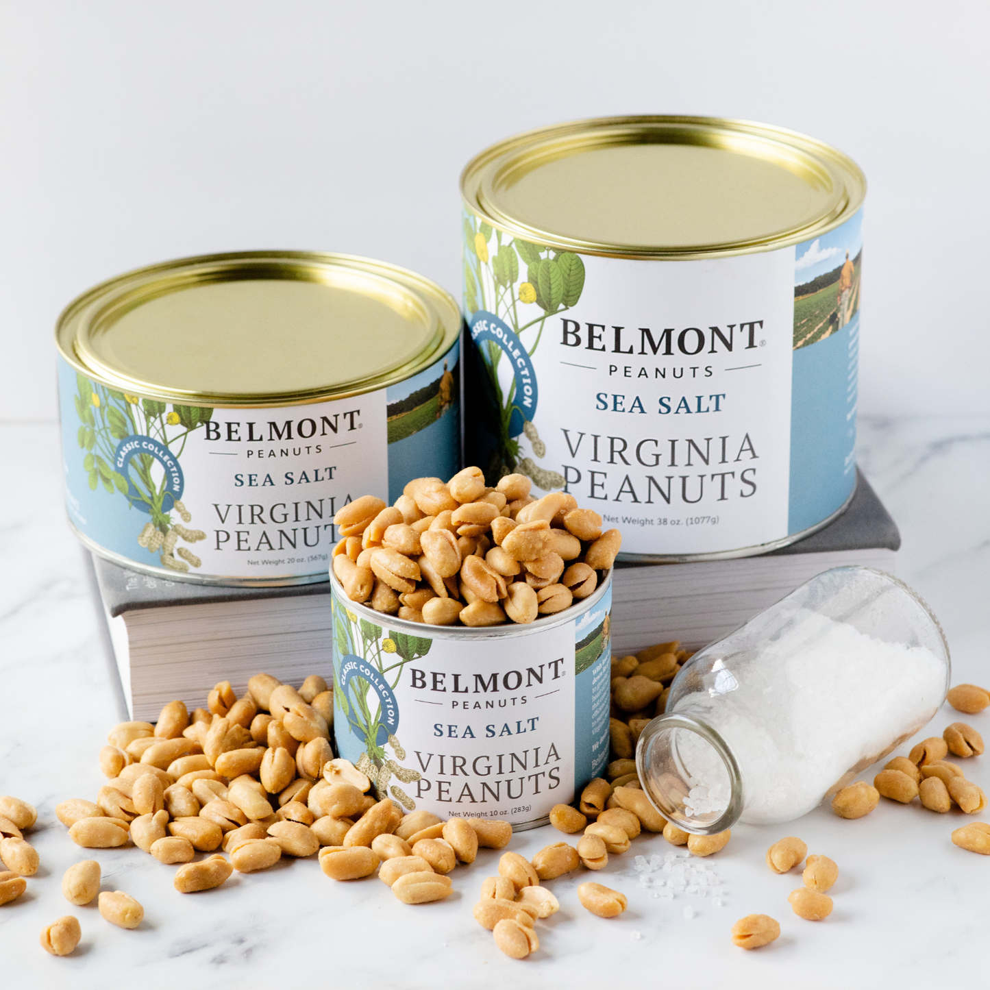 Three cans of Belmont Sea Salt, one open and filled with Virginia peanuts, are set on books beside a tipped jar of spilled Sea Salt, with loose peanuts scattered on a marble surface.