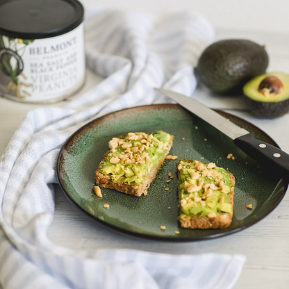 Two slices of avocado toast topped with chopped Virginia peanuts are on a green plate with a knife. In the background are a striped cloth, a can of Sea Salt & Black Pepper gourmet peanuts, and two avocados, one halved.