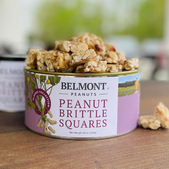 A round tin labeled Peanut Brittle Squares rests on a wooden surface, filled with chunky pieces of peanut brittle. The tin displays floral designs and peanut graphics, showcasing this classic sweet snack.