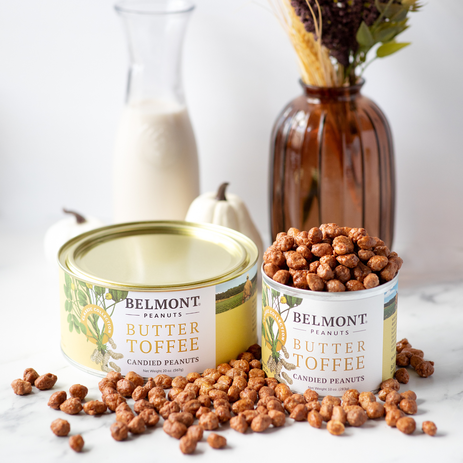 Two cans of Butter Toffee peanuts, one open and full, with buttery candy-coated peanuts spilling onto a marble surface. A vase of dried flowers and a bottle of milk are seen in the background.