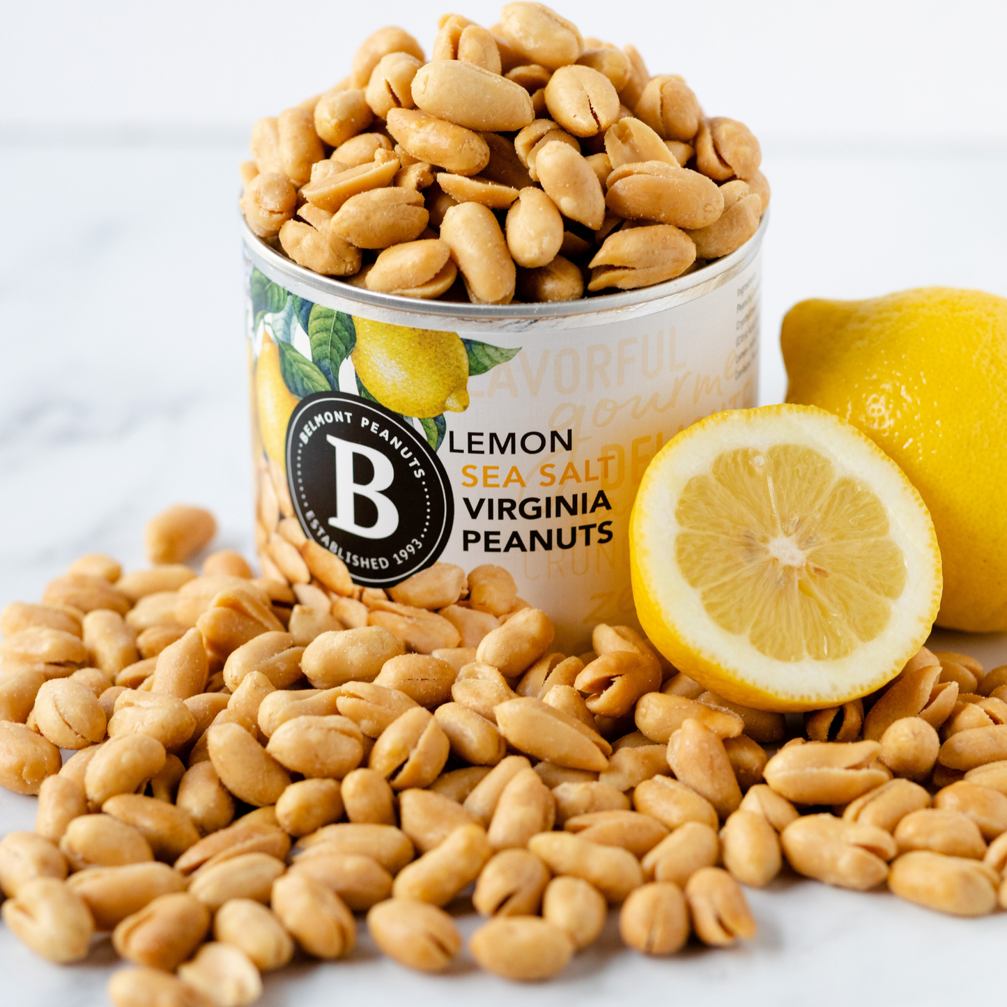 A can of Lemon Sea Salt Virginia Peanuts sits on a white surface, surrounded by scattered peanuts and a halved lemon. The can displays a lemon illustration and the Belmont Peanuts logo.