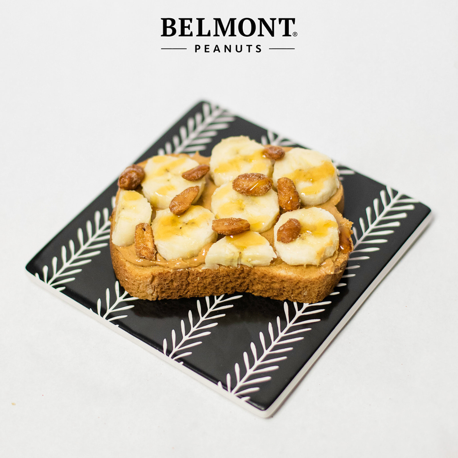 A slice of toast with banana, Honey Roasted peanuts, and honey is served on a black plate with a white leaf pattern. The BELMONT PEANUTS Honey Roasted logo is displayed above the plate on a white background.