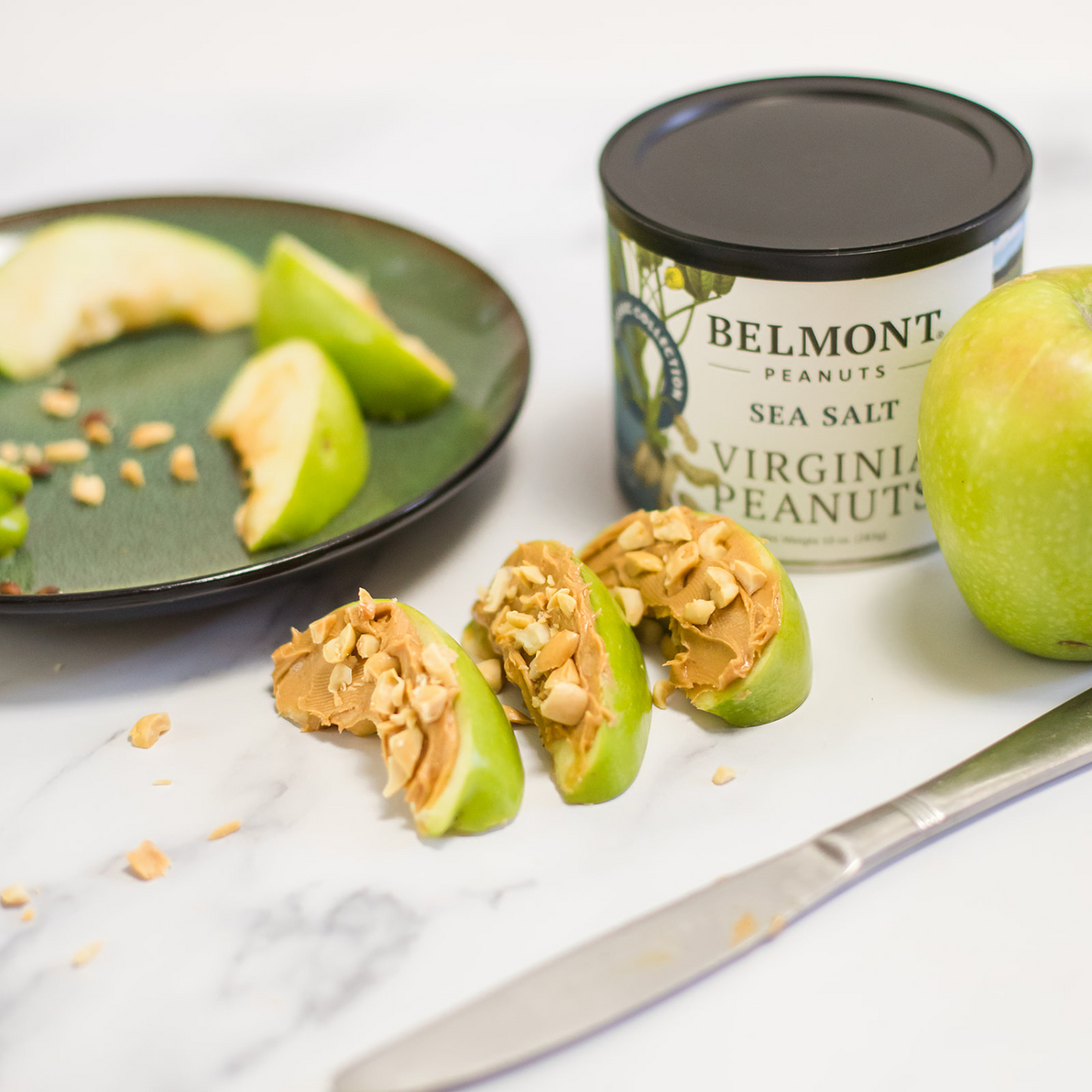 Sliced green apples with peanut butter and chopped Sea Salt peanuts, a knife, a whole green apple, and a can of Sea Salt rest on marble. A plate of more apple slices appears in the background.