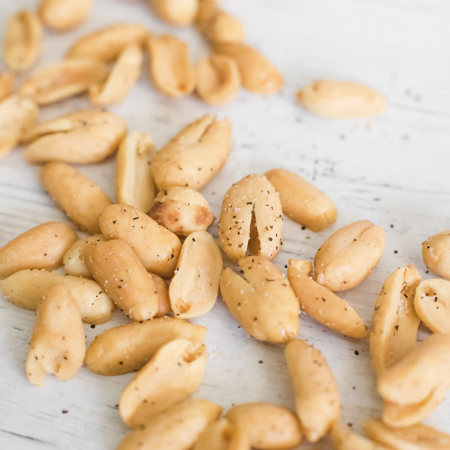 Sea Salt & Black Pepper peanuts roasted to perfection, scattered on a white wooden surface for a gourmet touch.
