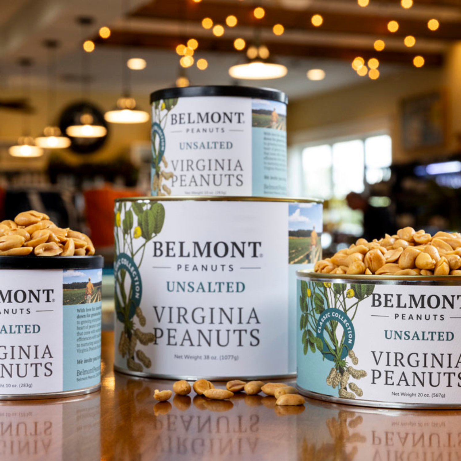 Three cans of Belmont premium peanuts, labeled Unsalted, are stacked on a wooden table with unshelled peanuts spilling out. Warm, blurred lights and a cozy shop interior create an inviting background.