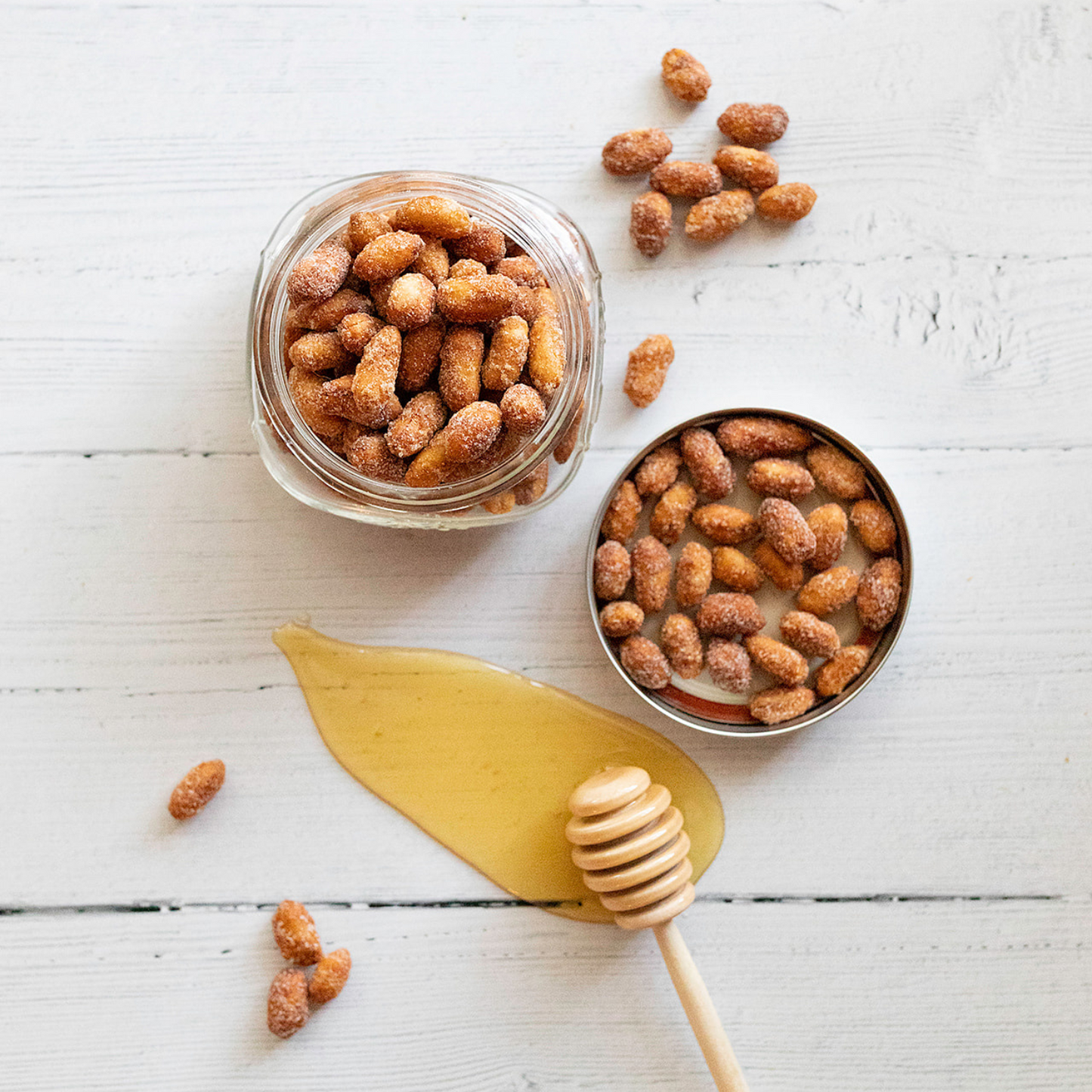 A glass jar with a metal lid, filled with Honey Roasted sugared almonds, sits on a white wooden surface. Nearby are gourmet peanuts and a honey dipper resting in honey, with scattered almonds around.