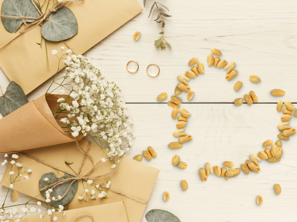 The Anniversary bouquet of white baby’s breath flowers, tied with twine, is displayed on kraft paper envelopes next to two gold rings and a yellow seed heart on a white wooden surface.