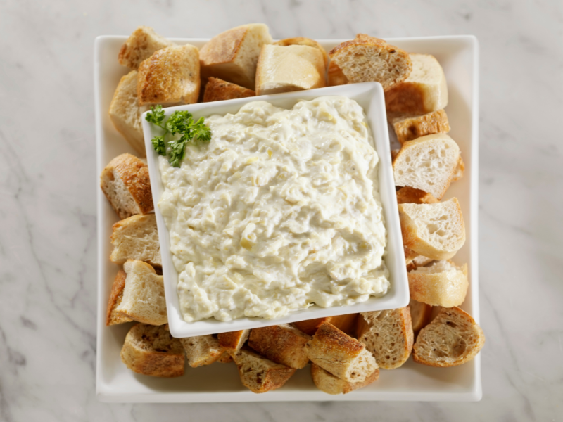 A square white bowl of Artichoke Dippin' Sauce, garnished with parsley, rests on a matching plate surrounded by baguette slices atop a marble surface.