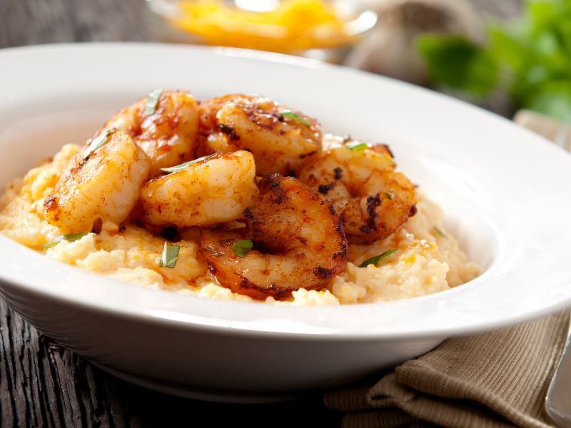 Shrimp & Grits features creamy stone-ground grits topped with seasoned sautéed shrimp and fresh green onions—Southern comfort served in a white bowl on a rustic wooden table.