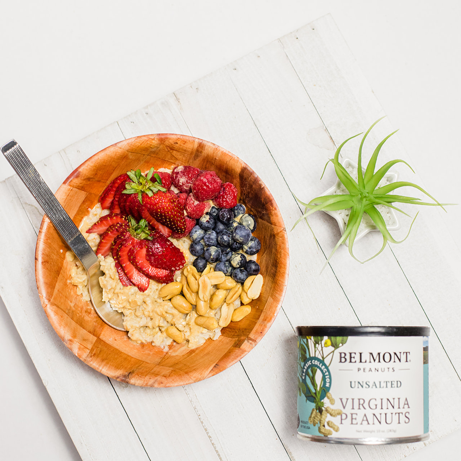 A wooden bowl with oatmeal topped with strawberries, blueberries, and Unsalted peanuts sits beside a small potted air plant and a can of Unsalted on a white wooden surface.
