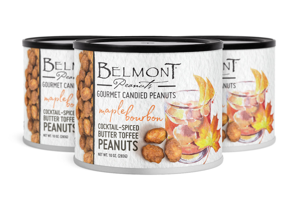 Three cans of Butter Toffee Maple Bourbon peanuts are shown. The white cans display an illustration of a cocktail, ice, maple leaves, and peanuts, with text emphasizing their sweet taste and cocktail-inspired spice.
