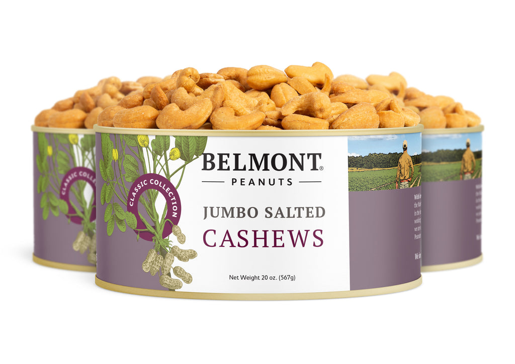 Three tins of Jumbo Salted Cashews are pictured, with roasted cashews spilling out. The purple and white label displays nut illustrations and farming scenes, emphasizing this healthy snack.