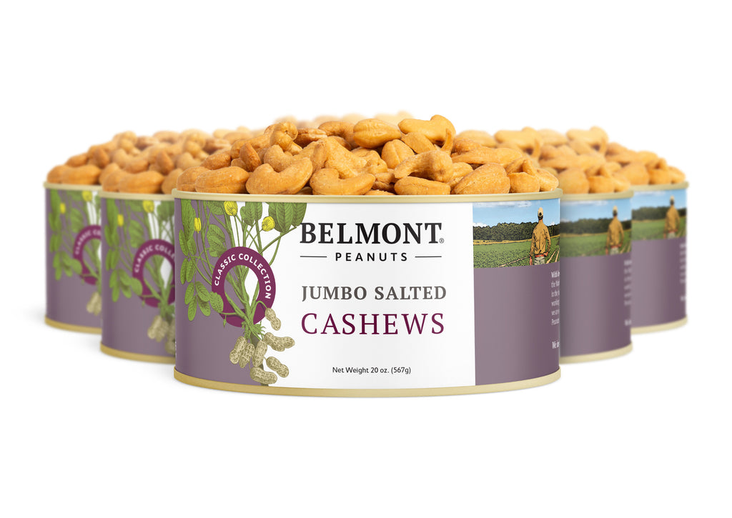 Four cans of Jumbo Salted Cashews are lined up, each with a purple and white label showing roasted cashews and a farm scene, emphasizing these whole cashews as a tasty, healthy snack.