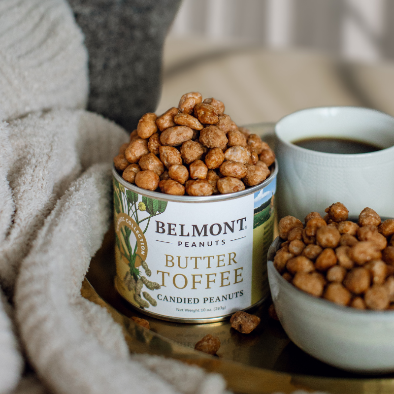 Can of Belmont Butter Toffee peanuts on a table with a cup of coffee and more peanuts.