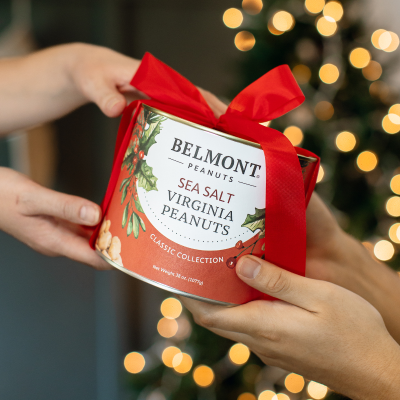 Belmont Peanuts sea salt Virginia peanuts with a red gift ribbon being handed over with a blurred Christmas tree in the background.
