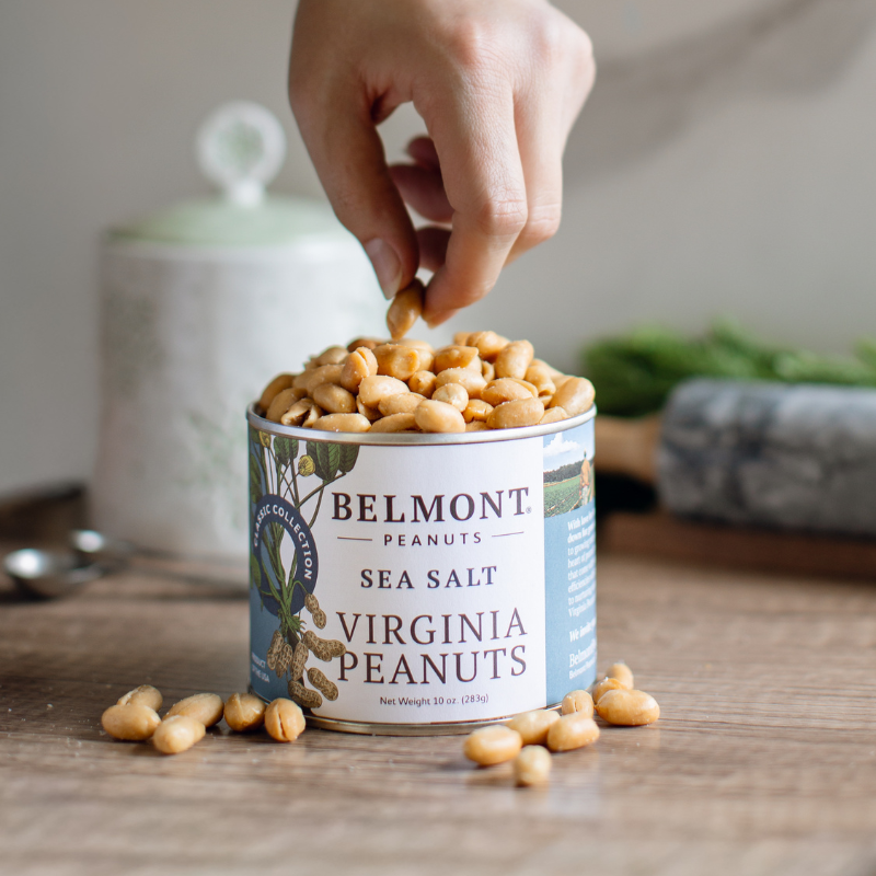 Hand picking peanuts from a can of Belmont Virginia Peanuts on a wooden surface.